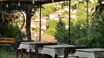 Un área de estar al aire libre con mesas y sillas, una pérgola con luces colgantes y vistas a una colina con edificios.