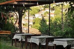 An outdoor seating area with tables and chairs, a pergola with string lights, and a view of a hillside with buildings.