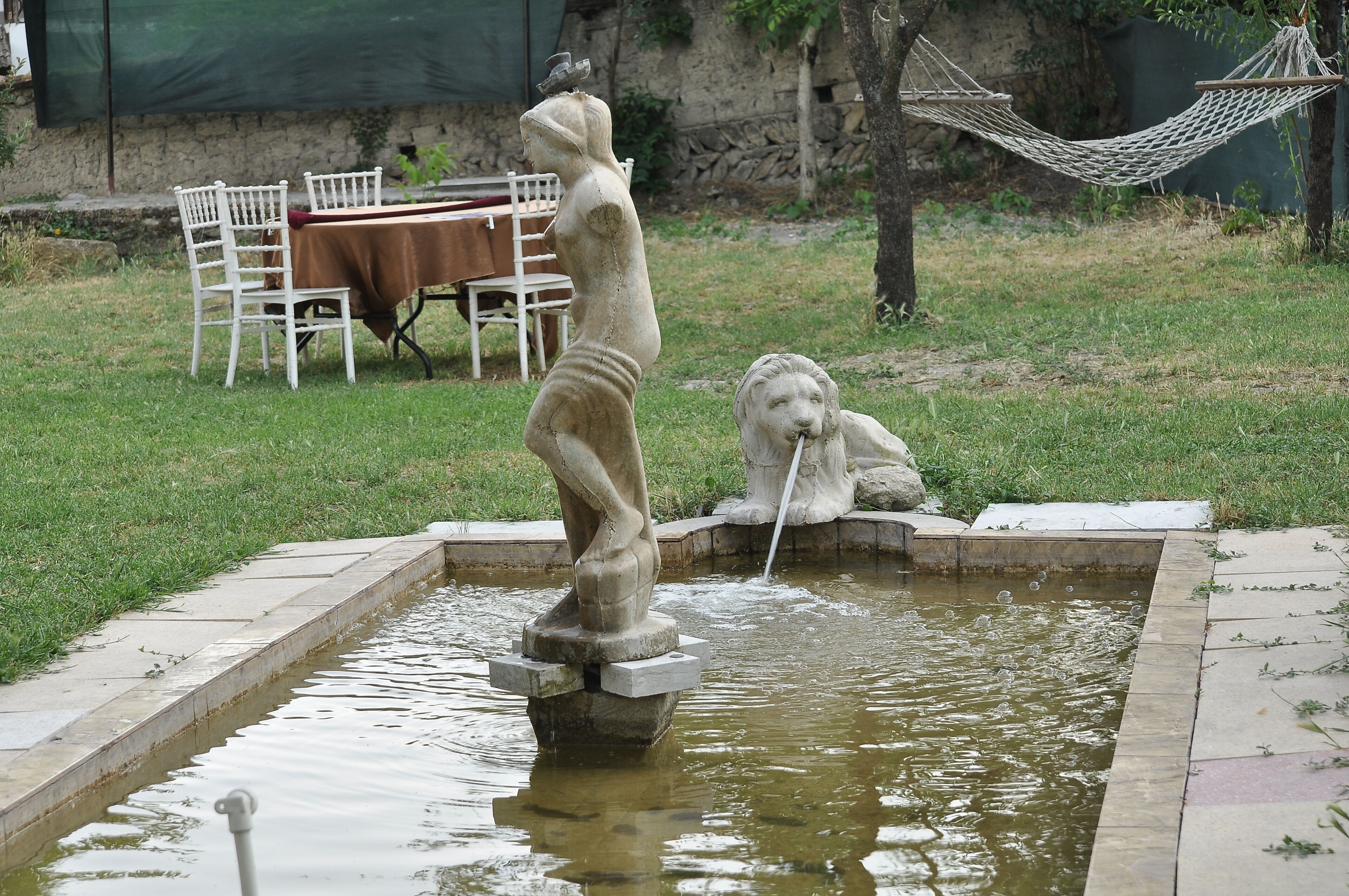 Una fuente con una estatua de una mujer y un león, rodeada de césped, una hamaca y una mesa con sillas.