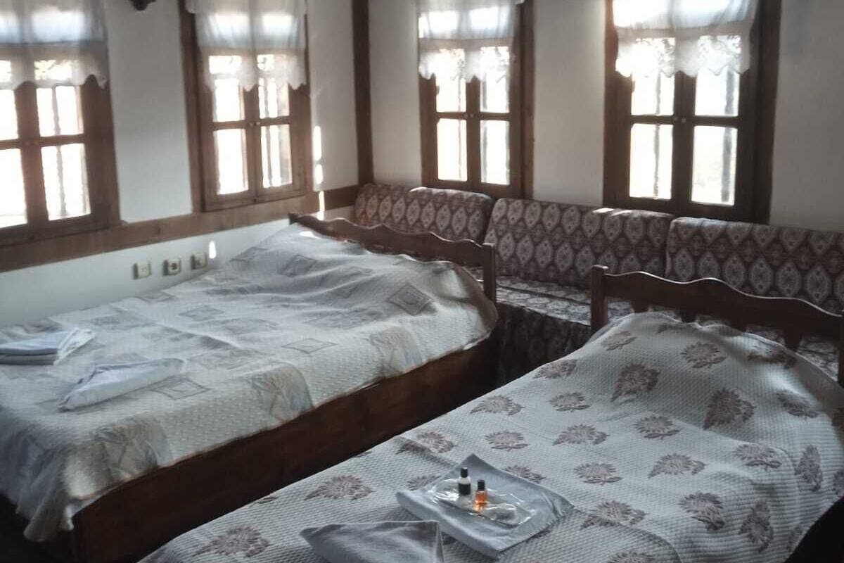 Quarto com duas camas, cada uma com colcha estampada e um bandejão com artigos de higiene. Há janelas com cortinas, vigas de madeira no teto e um painel de parede estampado.