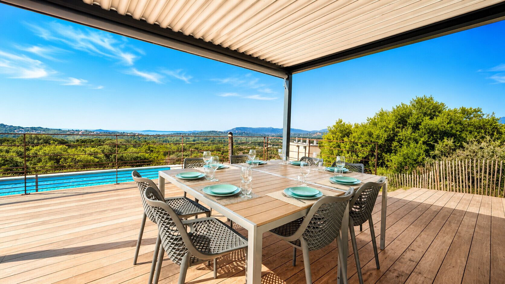 A wooden deck with a table set for dining, overlooking a pool and a scenic landscape.