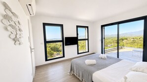 A bedroom with a bed, two large windows, a flat-screen TV, and a balcony with a view.
