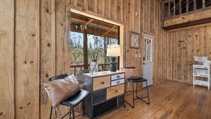 A wooden cabin interior with a desk, two chairs, a lamp, and a view through a large window.