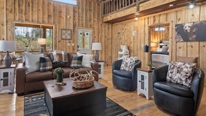 A rustic cabin interior with wooden walls, a brown leather sofa, two armchairs, a coffee table, and a dining area with a table and chairs.