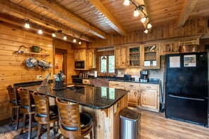 A wooden cabin interior with a kitchen featuring a central island, bar stools, and a black refrigerator. - Heavens Ridge-3-bedroom Pet Friendly Luxury Log Cabin Rental, Mountain Views, 30 Minutes to Great Smokey Mountains, High Speed Internet, 3 Decks, Hot Tub in Nantahala NC (Topton)