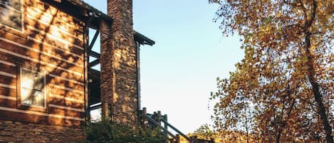 A rustic building with a stone chimney, wooden staircase, and a view of autumn trees.
