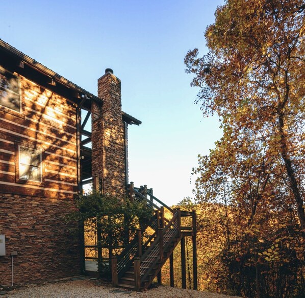 A rustic building with a stone chimney, wooden staircase, and a view of autumn trees.
