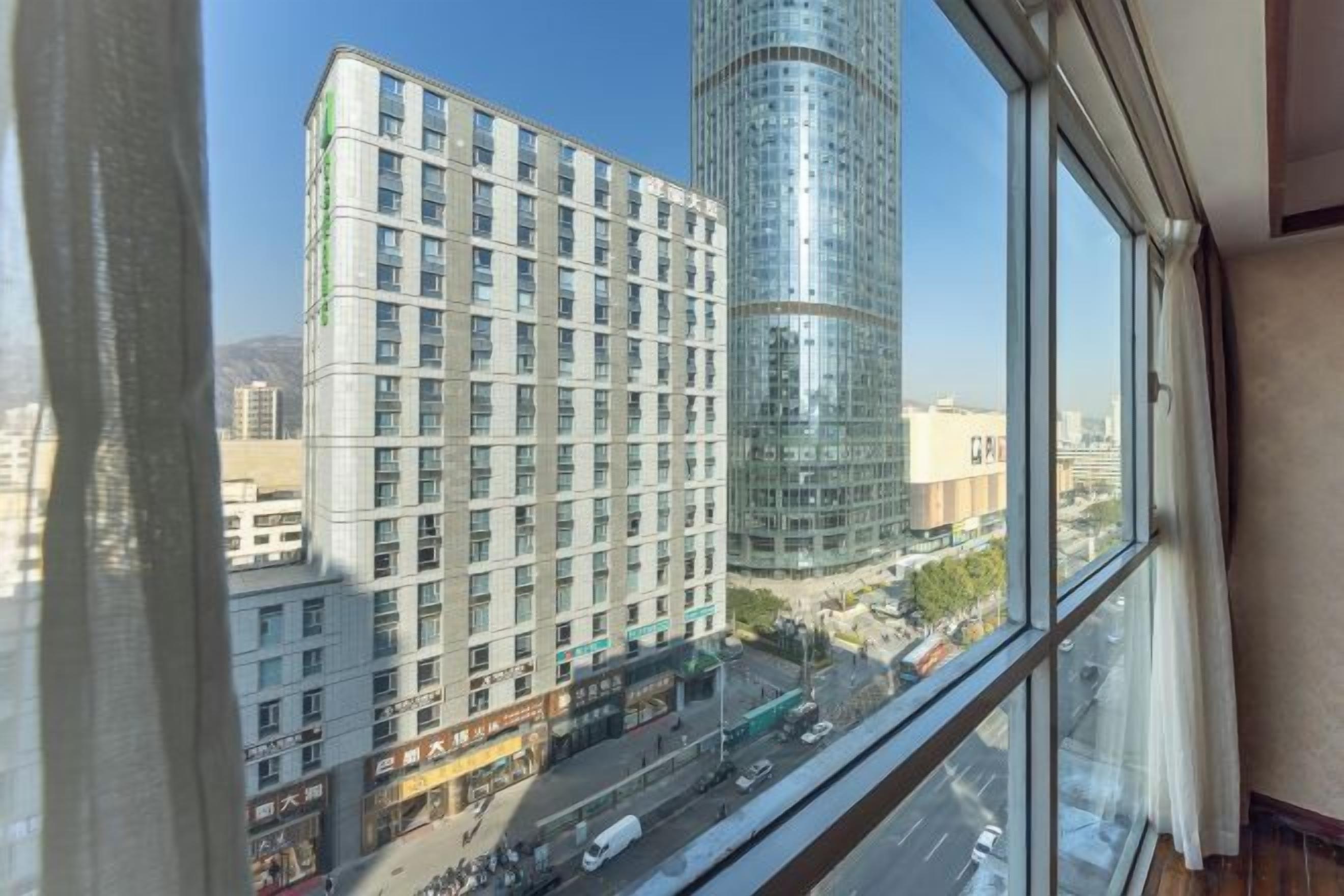 Une vue depuis une fenêtre donnant sur un paysage urbain, avec des bâtiments modernes, un ciel dégagé et une rue animée par la circulation.