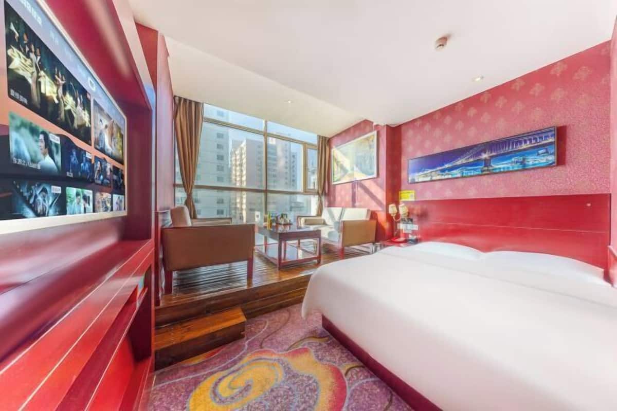 A hotel room with a large bed, a desk, a chair, a TV mounted on the wall, and a window with a city view.
