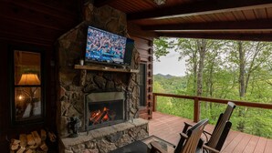 Une cheminée en pierre avec un téléviseur fixé au-dessus, une terrasse en bois avec des sièges d’extérieur et une vue sur un paysage boisé.