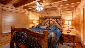 Une chambre dans une cabane en bois, avec un lit, deux tables de chevet, un ventilateur de plafond et une fenêtre avec des stores.