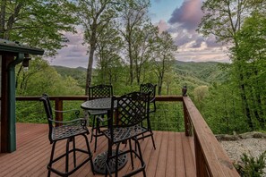 A wooden deck with metal chairs and a table, overlooking a forested valley.