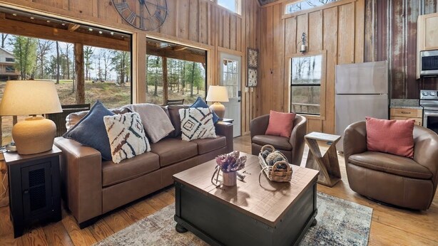 A cozy living room with a brown leather sofa, a coffee table, and a fireplace. There are large windows offering a view of the outdoors.