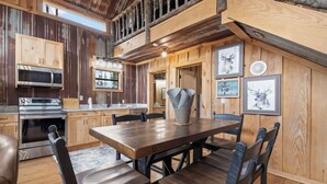 A rustic cabin interior with a wooden dining table, chairs, a kitchen with a microwave and oven, and framed deer artwork on the walls.