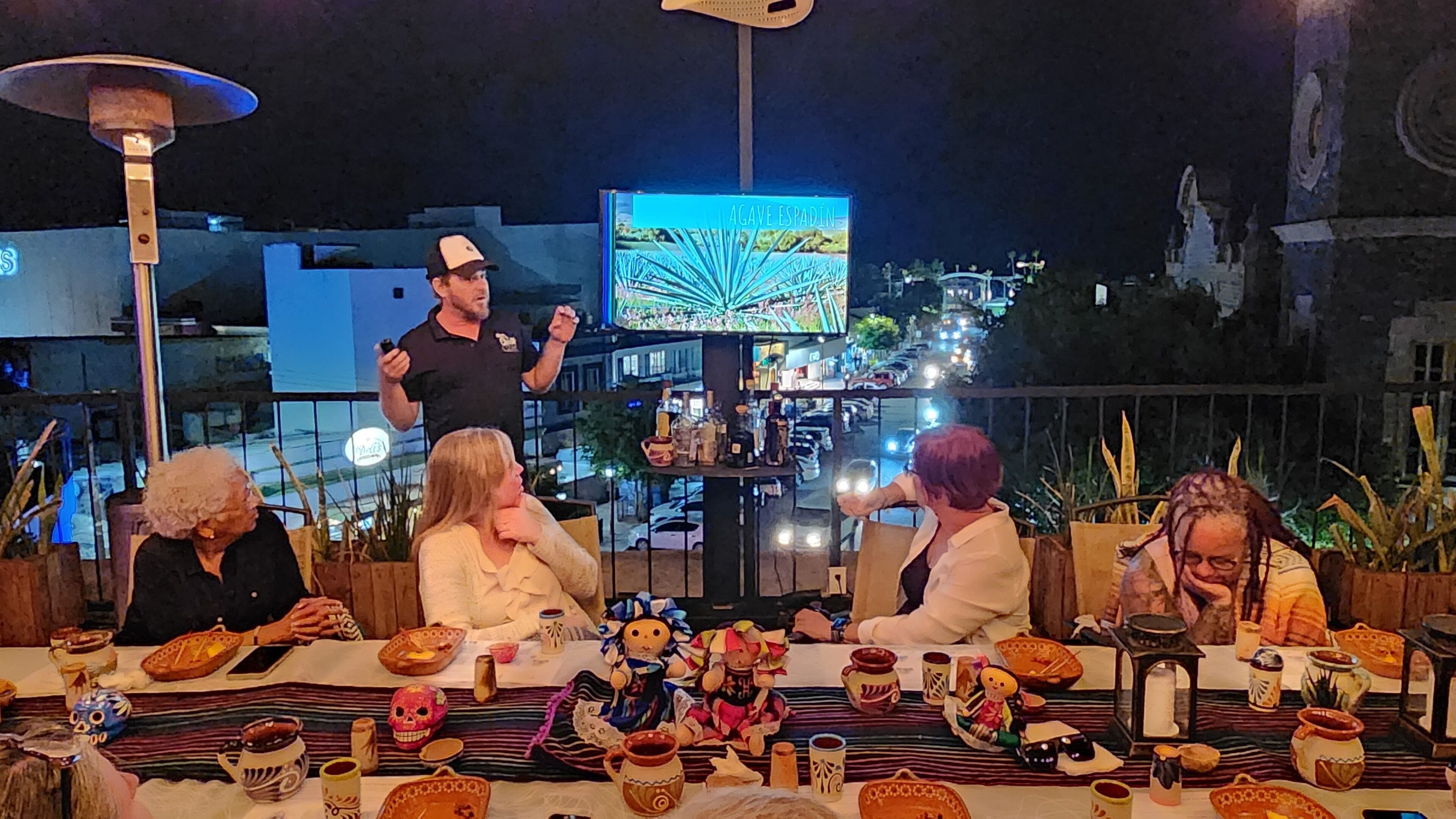 Um grupo de pessoas jantando ao ar livre à noite, com um horizonte urbano ao fundo.