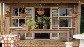 A wooden deck with a table and chairs, a hanging basket with flowers, and a view of the countryside.