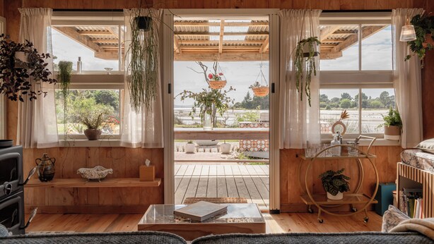 A cozy living room with wooden flooring, a sofa, a coffee table, and a view of a garden through large windows.