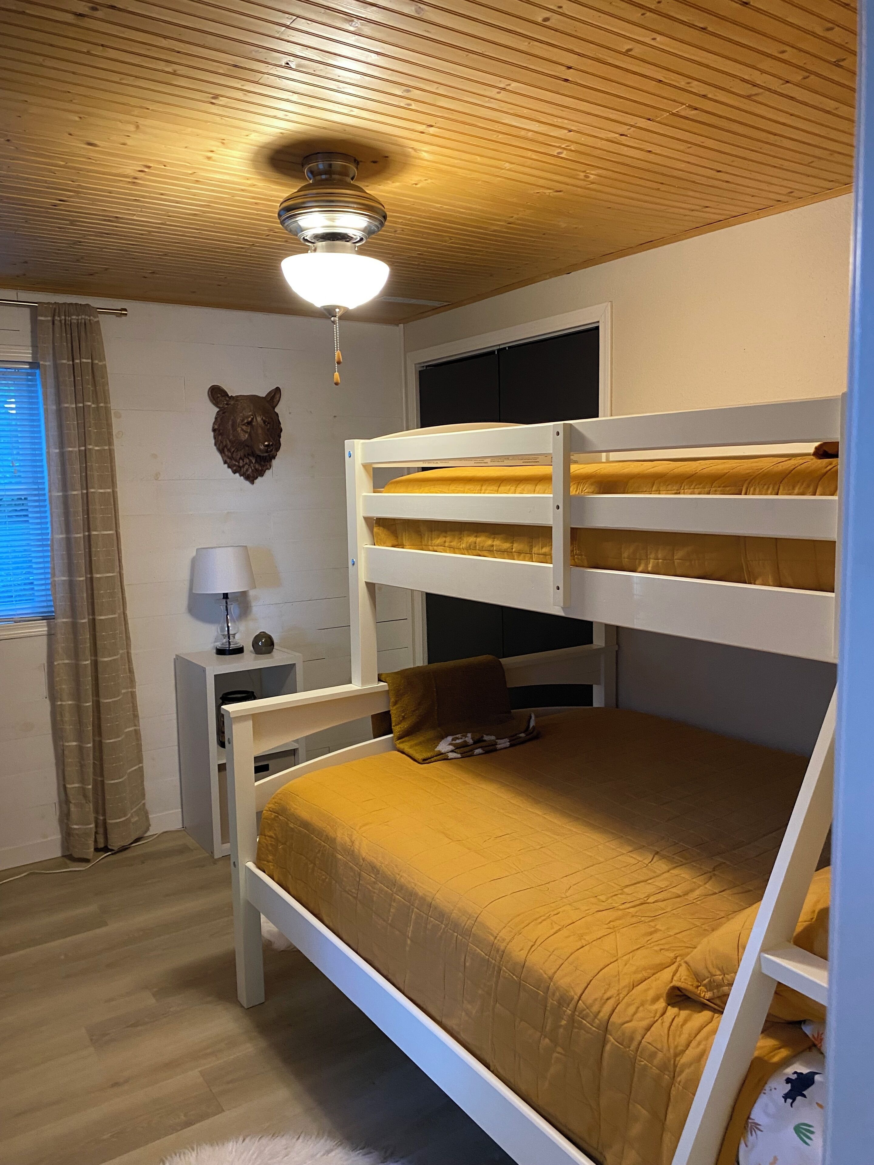 A bunk bed room with a ceiling fan, a wall-mounted bear head decoration, a bedside table with a lamp, and a window with curtains.