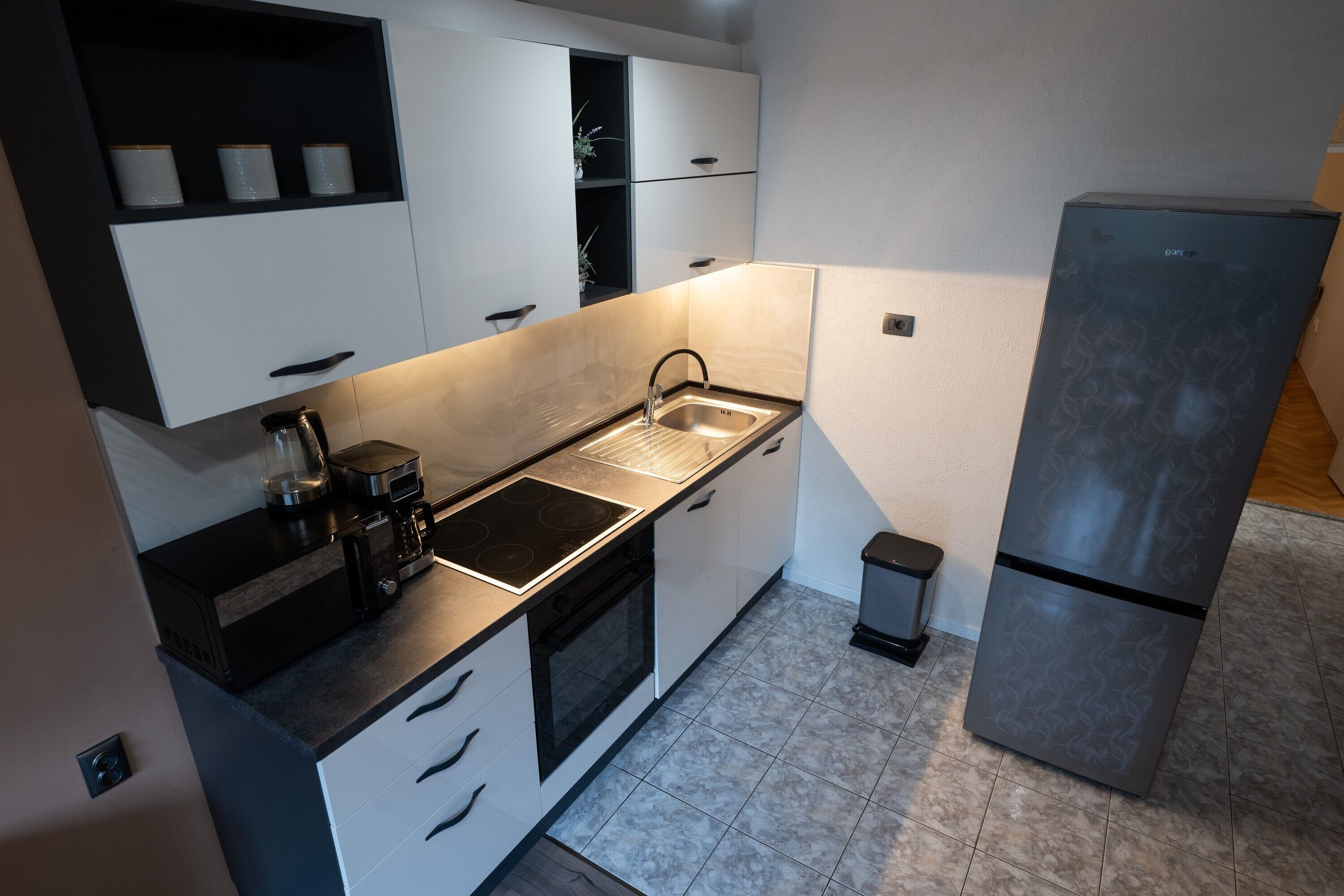 A modern kitchen with white cabinets, a black countertop, a stainless steel sink, a built-in oven, and a refrigerator.