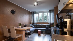 Eine moderne Küche mit Einbauherd, Kaffeemaschine und Kühlschrank. Es gibt eine Essecke mit Tisch und Stühlen, sowie eine Couch im Hintergrund.