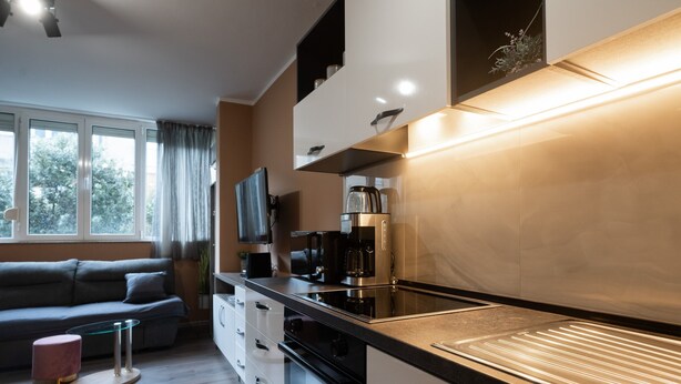 Eine moderne Küche mit Einbauofen, Kaffeemaschine und Fernseher. Im Wohnbereich befindet sich eine Couch und ein kleiner runder Tisch.