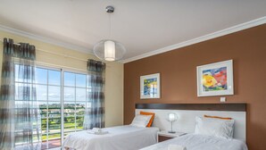 Hotellihuone, jossa on kaksi sänkyä, suuri ikkuna verhoilla, kattovalaisin ja kaksi kehystettyä taideteosta seinällä.