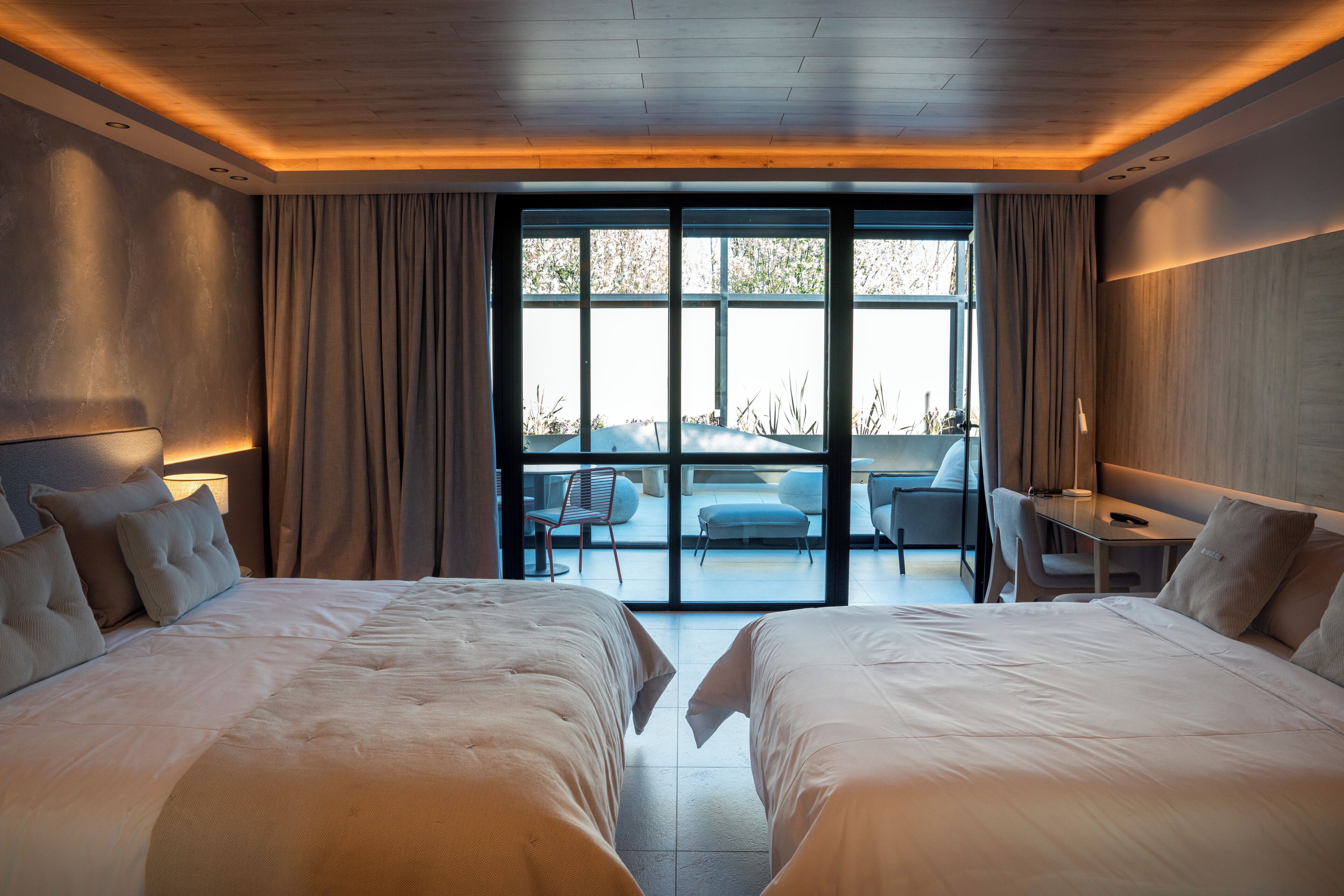A modern hotel room with two large beds, a desk, and a view of the outdoors through large windows.