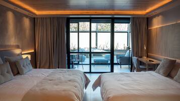 A modern hotel room with two large beds, a desk, and a view of the outdoors through large windows.