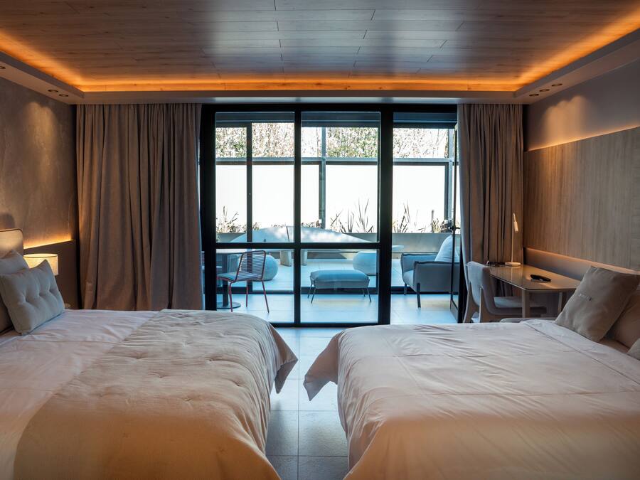 A modern hotel room with two large beds, a desk, and a view of the outdoors through large windows.