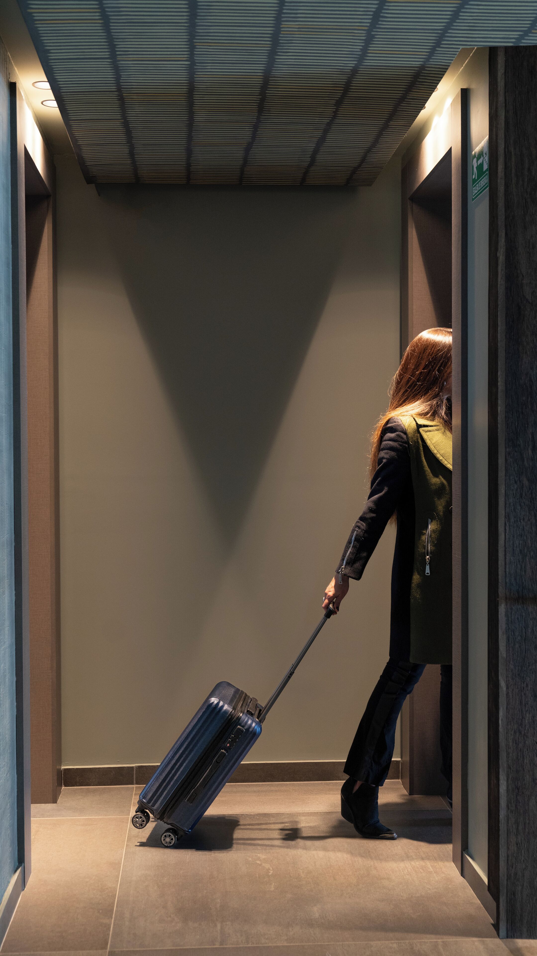 A person pulling a suitcase through a narrow hallway with a ceiling that has a grid pattern.