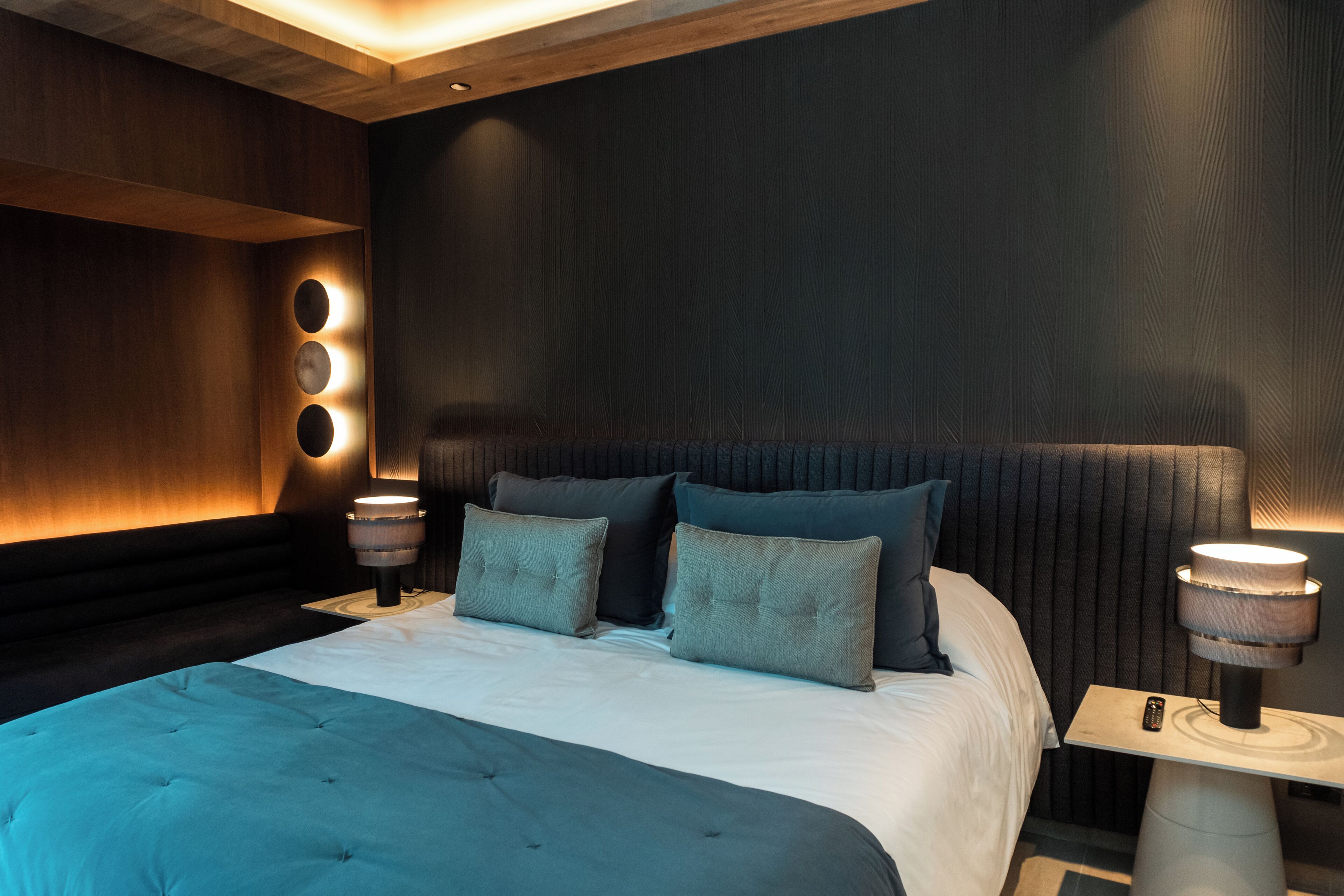 A modern bedroom with a large bed, dark wood paneling, and three bedside lamps.
