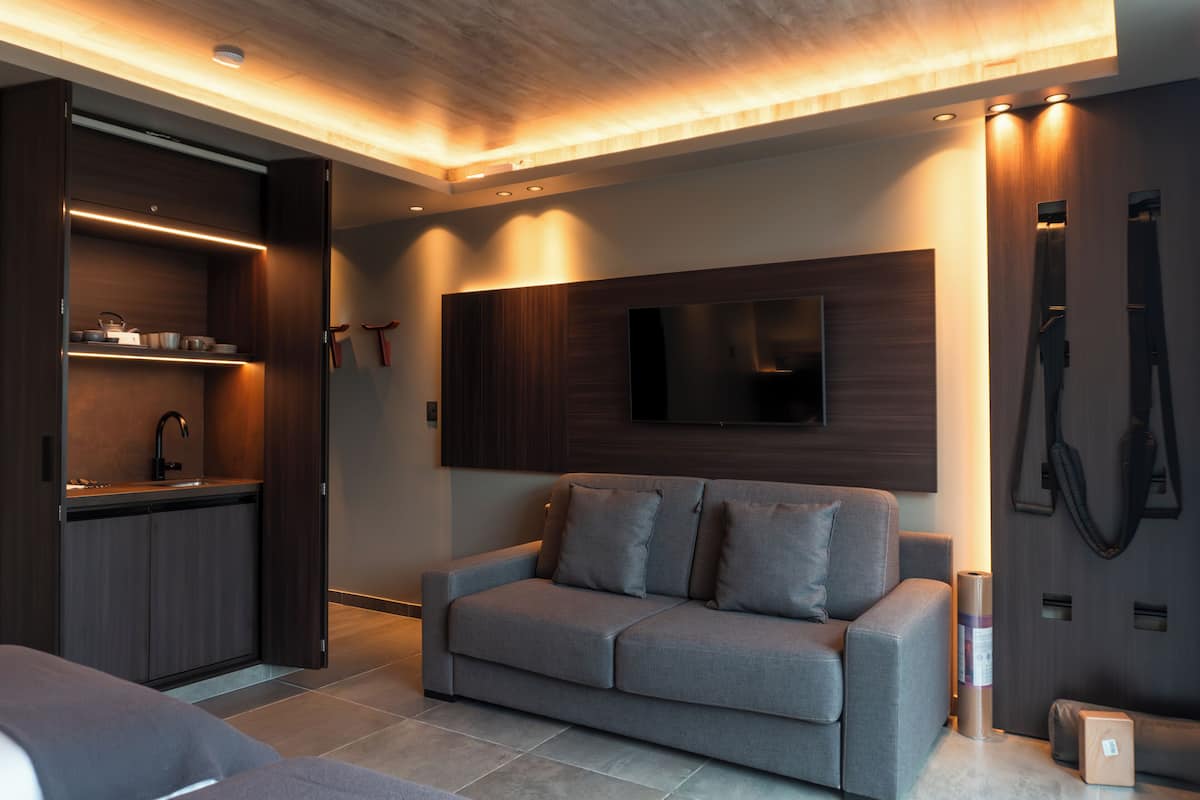 A modern living room with a sofa, a flat-screen TV mounted on the wall, and a built-in kitchen area with a sink and cabinets.