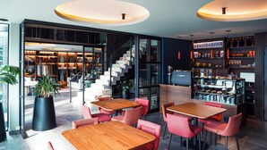 Un intérieur moderne avec des tables en bois et des chaises roses, un escalier et un coin bar avec des étagères remplies de bouteilles.