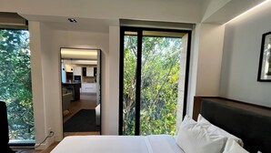 A modern hotel room with a large bed, a view of the outdoors, and a kitchenette in the background.