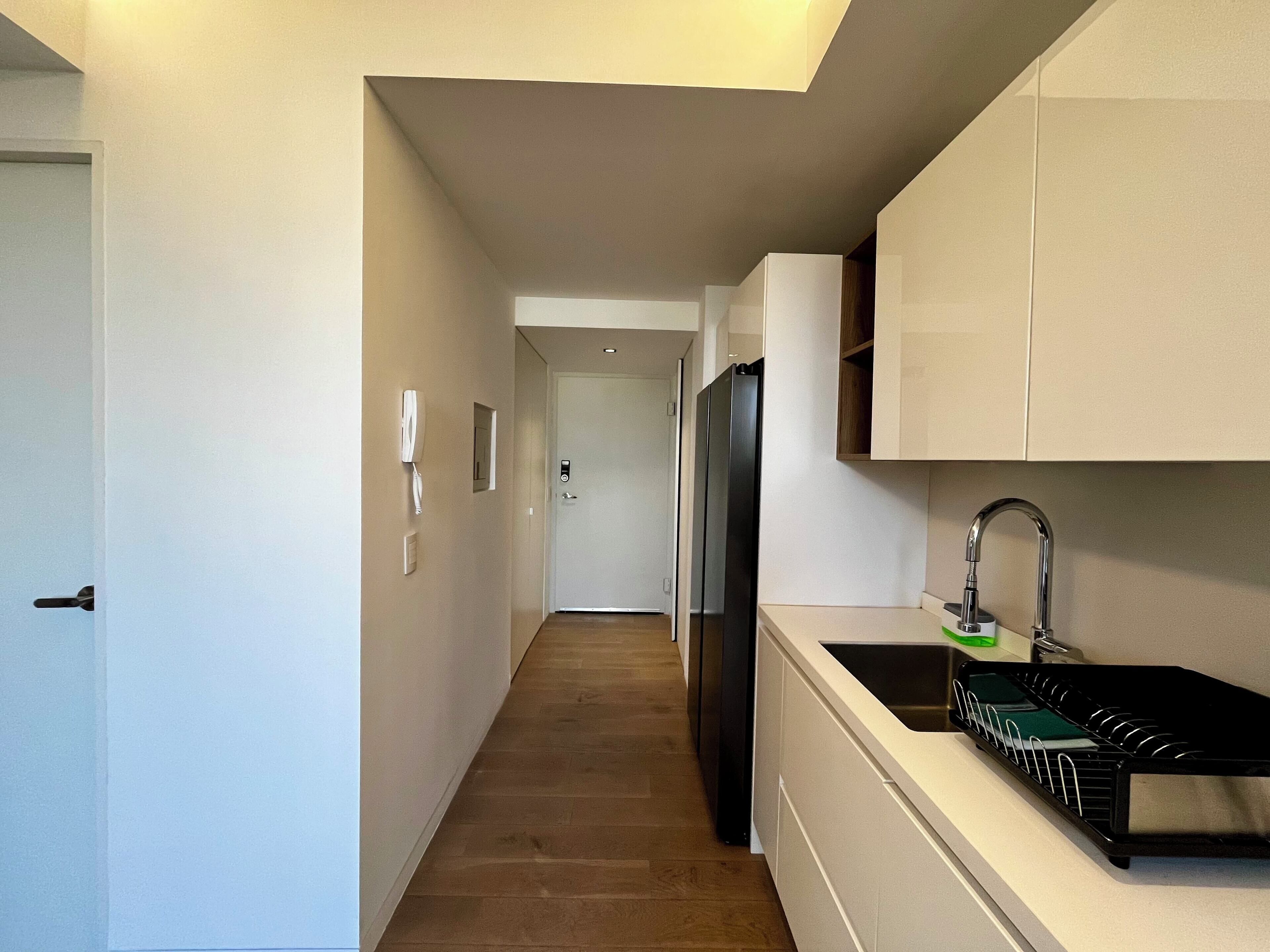 A narrow hallway leading to a kitchen with a refrigerator, a sink, and a dish rack.