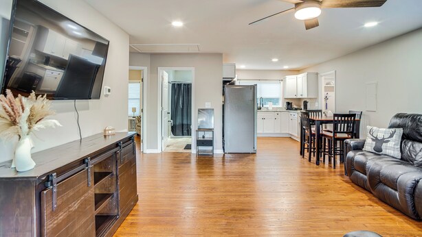 A spacious living area with a wooden floor, a leather sofa, a TV mounted on the wall, and a ceiling fan. - Home 14 Mi to Highpoint Health w/ Fenced Yard! (Hendersonville)