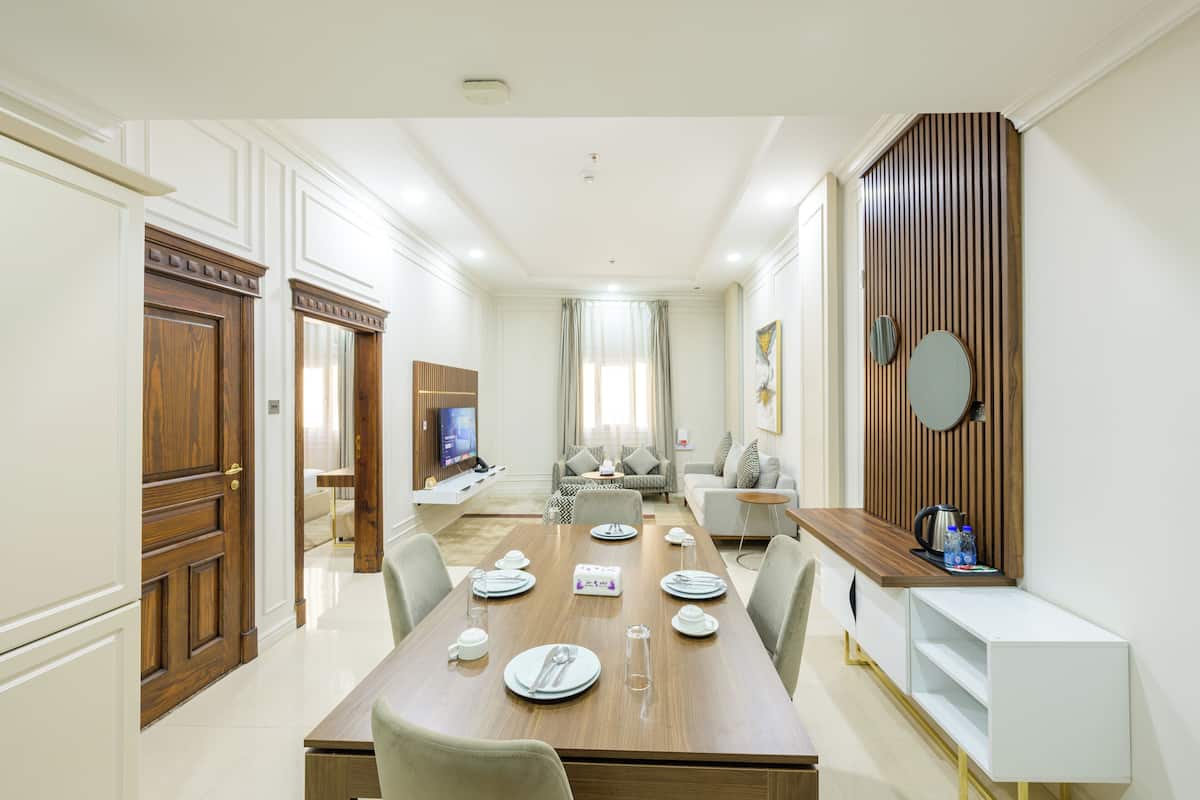 A modern dining area with a wooden table set for four, surrounded by chairs. There is a television in the background, a sofa, and a coffee table.