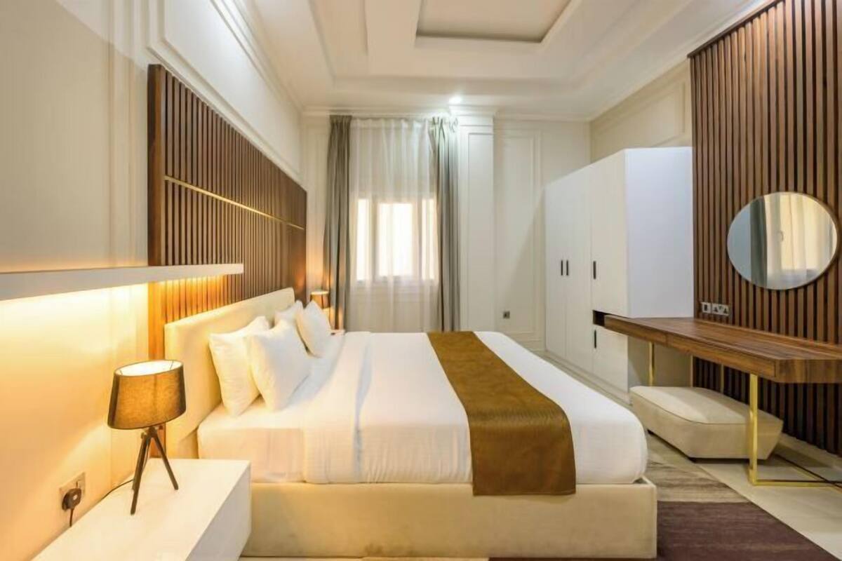 A hotel room with a bed, bedside table, lamp, wardrobe, mirror, and a view of the outdoors.