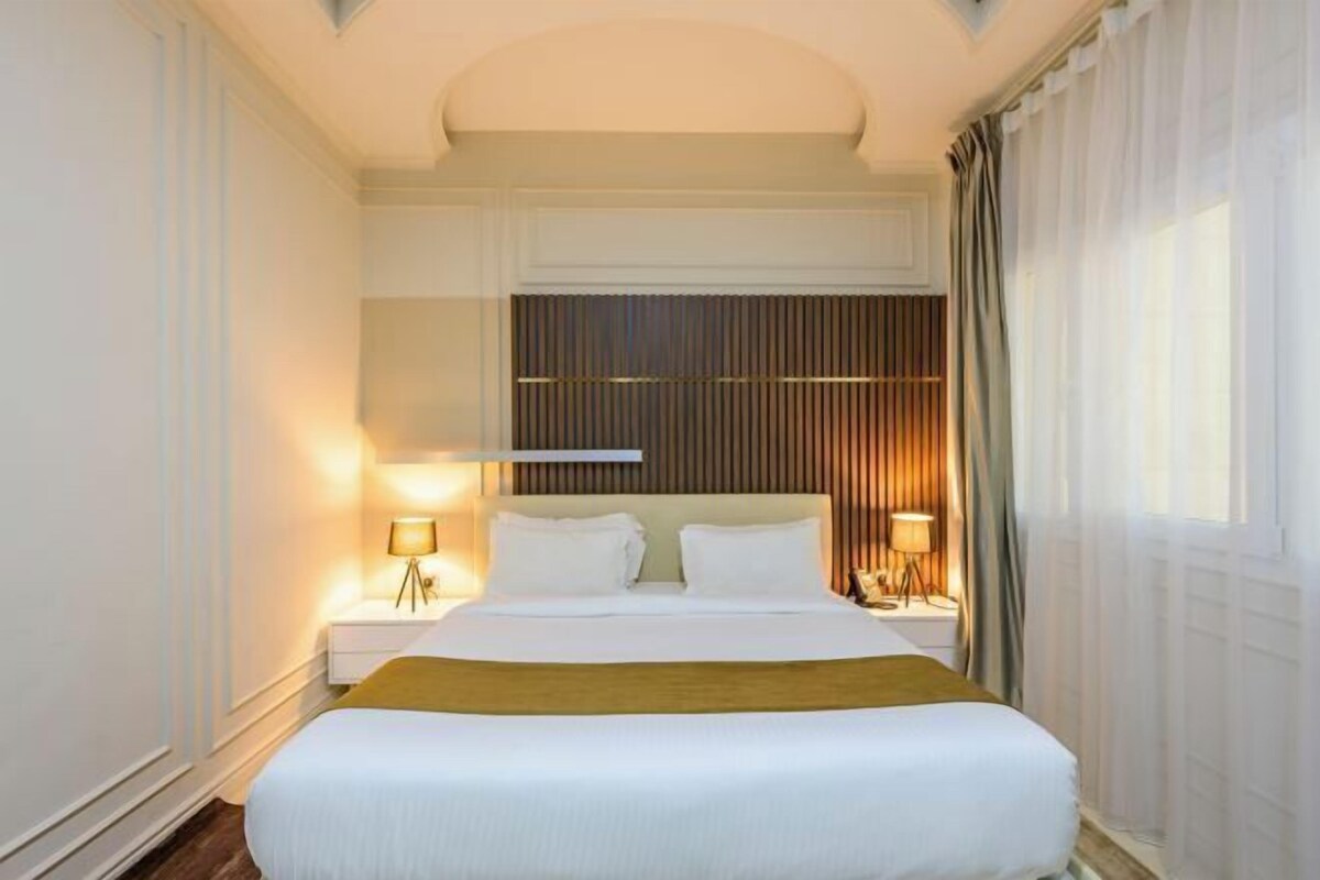 Ein Hotelzimmer mit einem großen Bett, zwei Nachttischen mit Lampen, einem Kopfteil mit vertikalen Lamellen und einem Fenster mit Vorhängen.