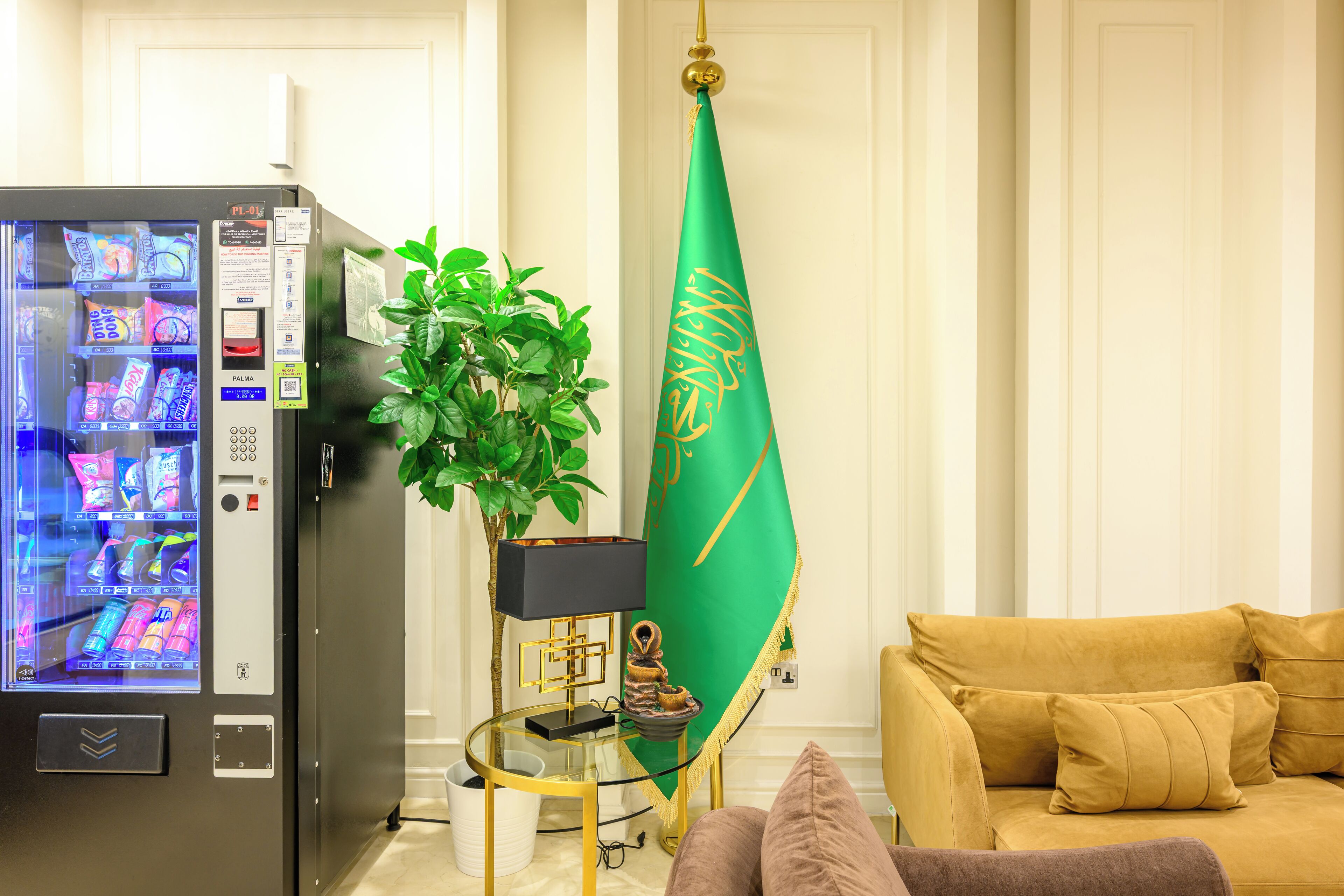 Uma sala de estar com máquina de vendas, uma bandeira verde, uma planta em vaso, uma mesa de centro de vidro e um sofá.