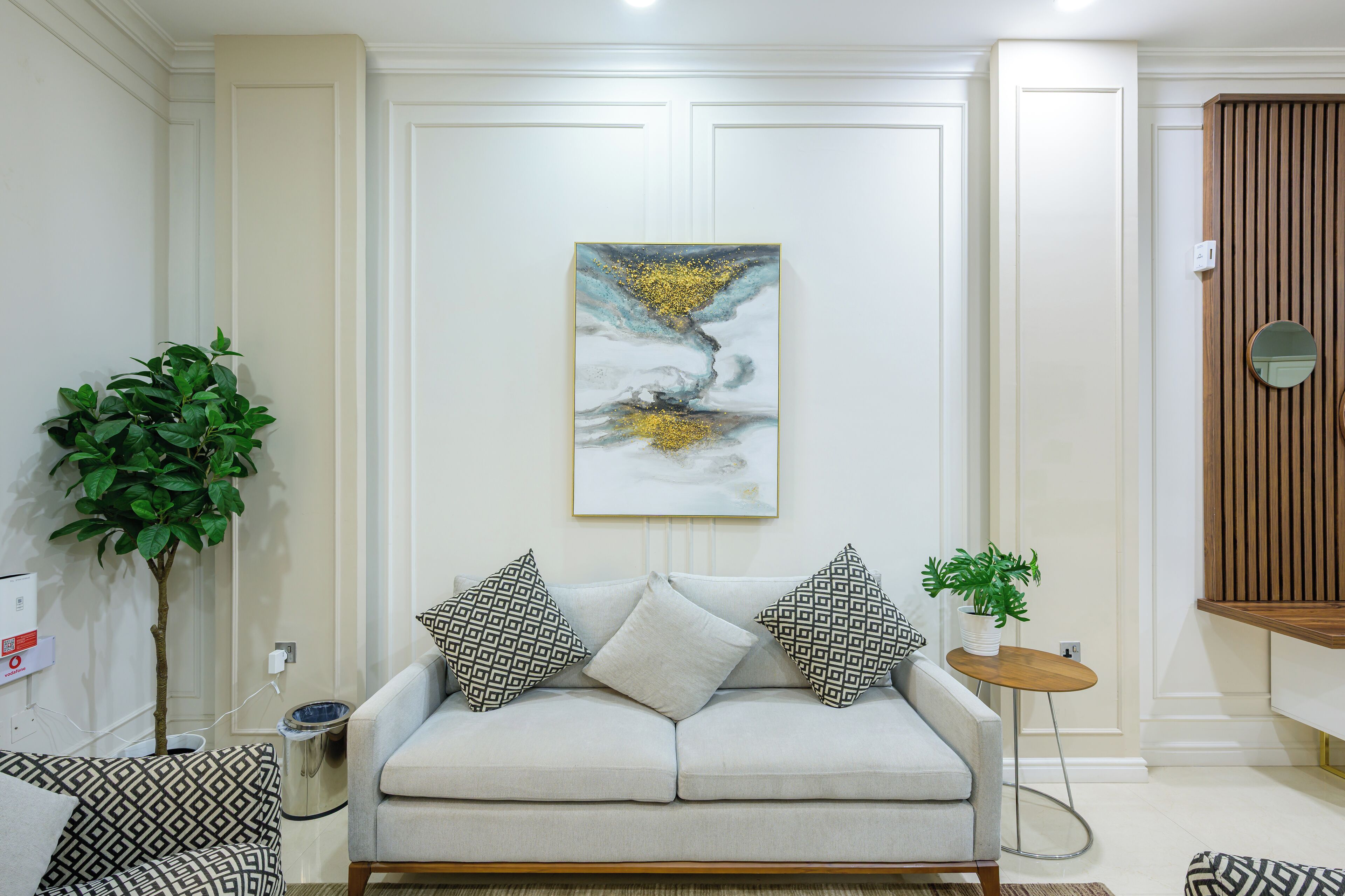 A living room with a light grey sofa, patterned cushions, a wall-mounted abstract painting, a small round table with a plant, and a potted plant beside the sofa.
