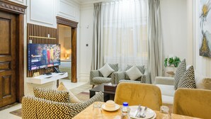 A well-lit living room with a television, patterned sofas, a dining table with cutlery, and a large window with curtains. - Abraj INN Royal Hotel (Doha)