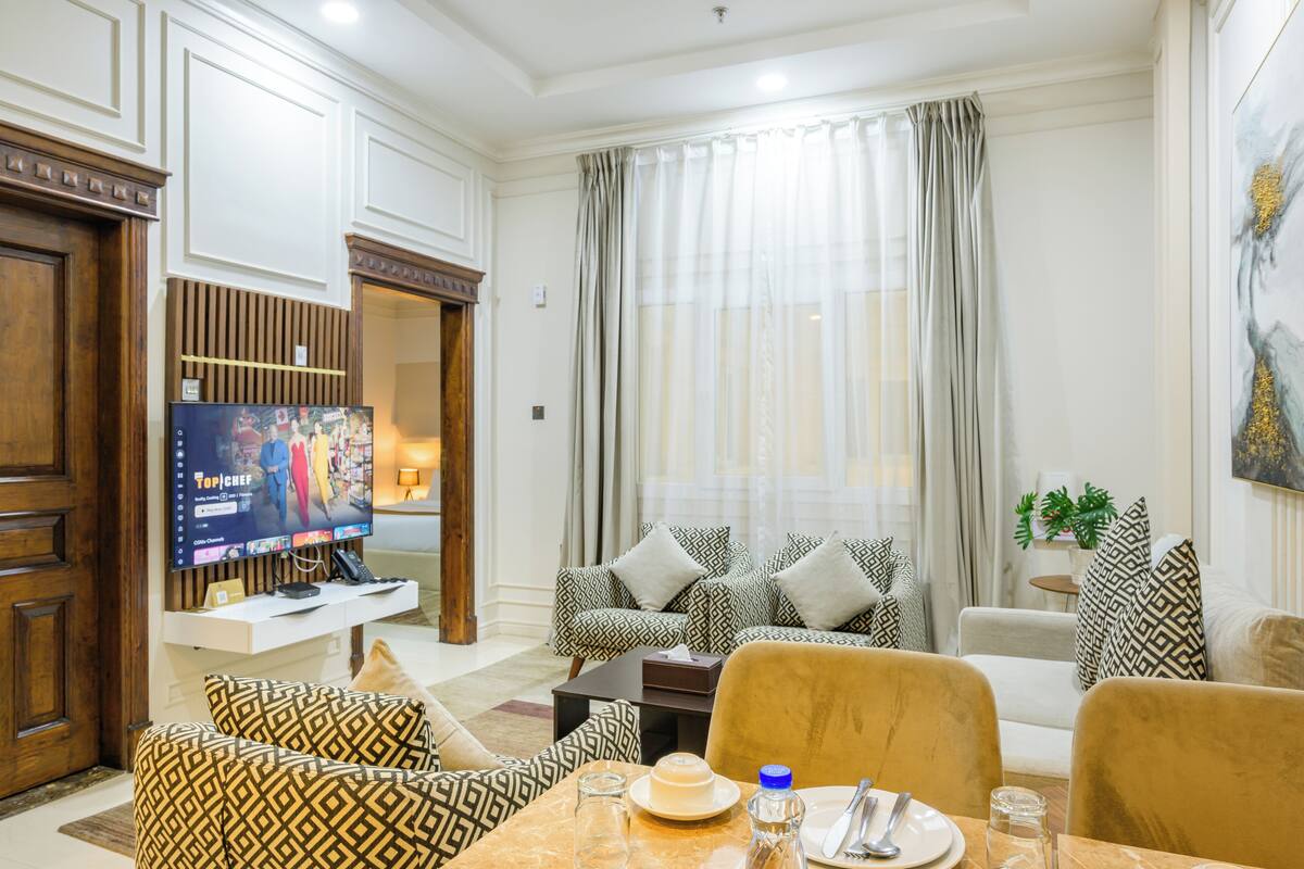 A well-lit living room with a television, patterned sofas, a dining table with cutlery, and a large window with curtains.
