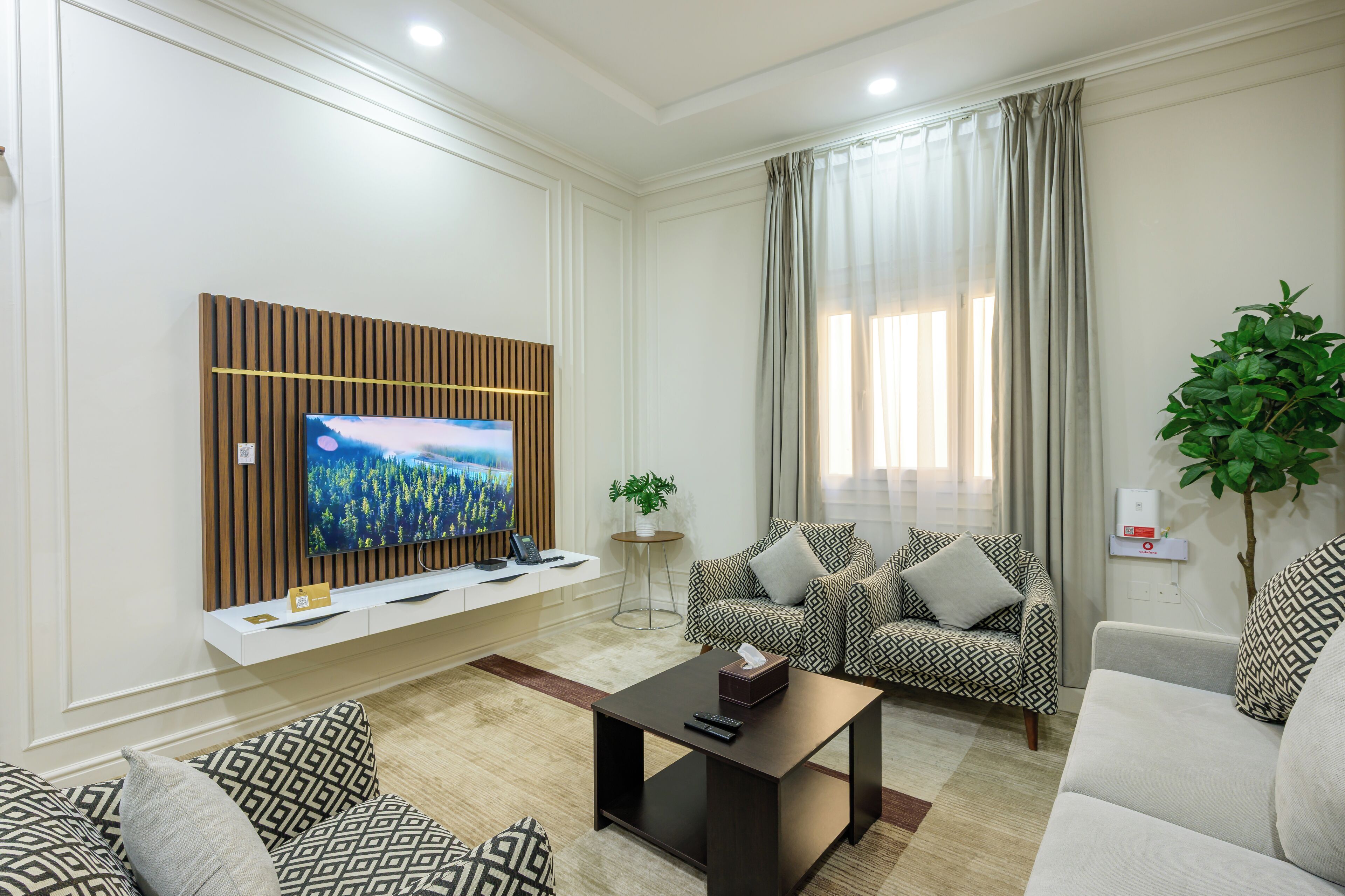 Een moderne woonkamer met een flatscreen televisie bevestigd aan een houten paneel, gestreepte banken, een salontafel en een potplant.