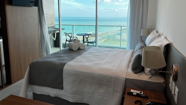 A hotel room with a large bed, a TV mounted on the wall, a balcony with a view of the sea, and a table with chairs outside. - RN STUDIO - APTO 620 (Maceio)