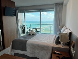 Une chambre d’hôtel avec un grand lit, un téléviseur fixé au mur, un balcon avec vue sur la mer et une table avec des chaises à l’extérieur.