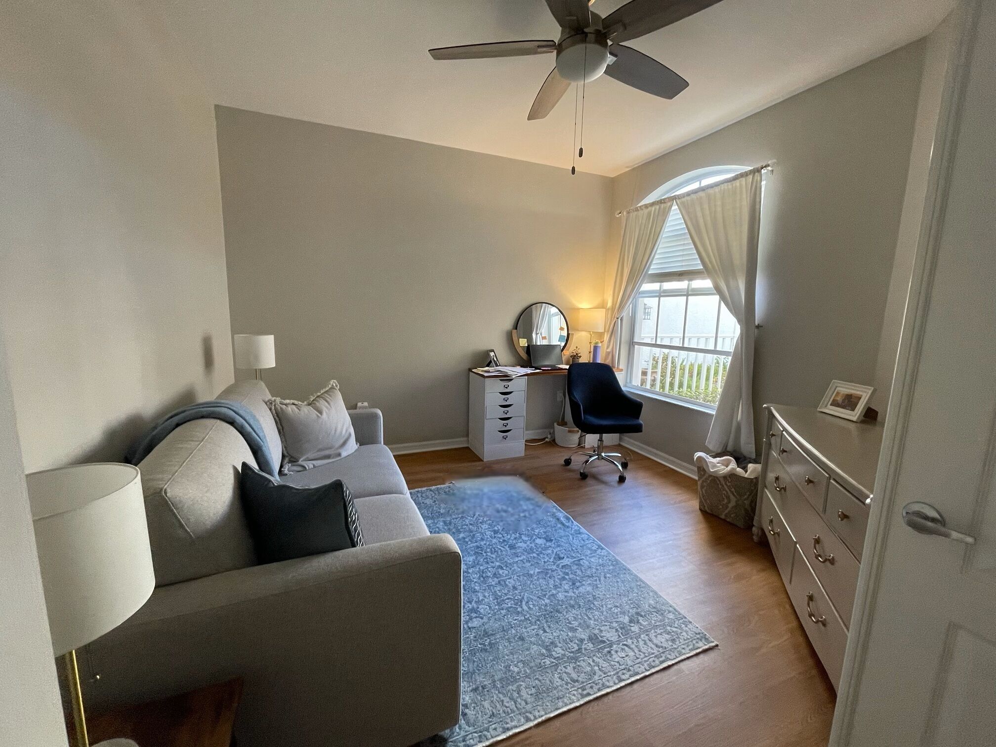 Ein Schlafzimmer mit einem Bett, einem Schreibtisch, einem Stuhl, einem Deckenventilator und einem Fenster mit Vorhängen.