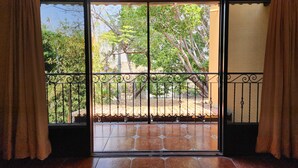 Vista através de uma porta de correr de vidro com portão de metal, para um pátio externo com piso de azulejos e vista de árvores e um edifício ao fundo.