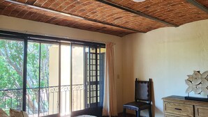 Quarto com teto de tijolos, uma cama grande com roupa de cama estampada, uma cadeira, cômoda e vista pela janela ampla.