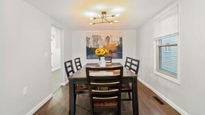 A dining area with a table and chairs, a cityscape painting on the wall, and a window with blinds.